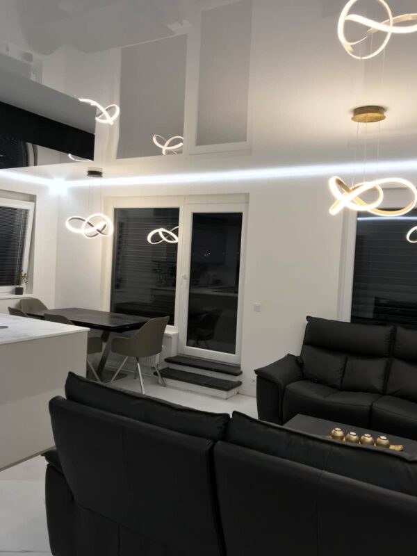 Moderní kuchyně a obývací pokoj s lesklým napínaným stropem, který dokonale odráží LED osvětlení a interiérový design  Luxusní černé sedačky a minimalistická kuchyň s ostrůvkem vytvářejí elegantní prostor 