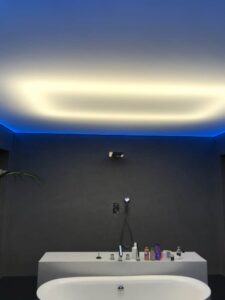 Napínaný strop s podsvícením a modrými LED prvky v moderní koupelně s volně stojící vanou a černými designovými detaily.