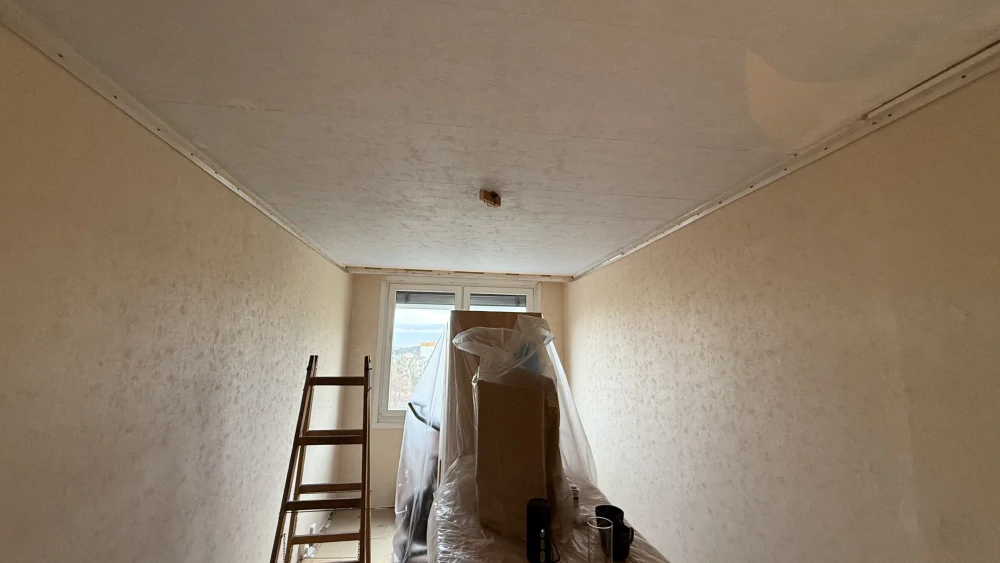 Pohled do místnosti s připraveným stropem pro montáž napínaného stropu, zakrytým nábytkem a hliníkovými lištami podél stěn.